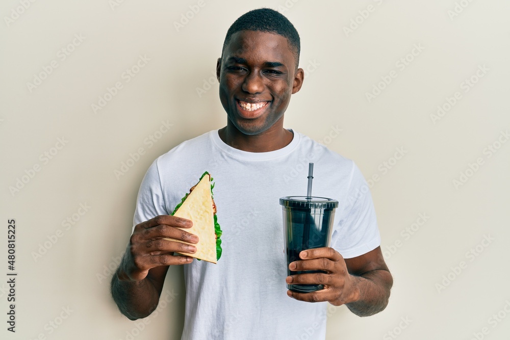 Young african american man eating sandwich and drinking soda smiling with a happy and cool smile on face. showing teeth. Young african american man eating sandwich and drinking soda smiling with a happy and cool smile on face. showing teeth.