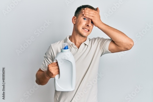 Young caucasian man holding detergent bottle stressed and frustrated with hand on head, surprised and angry face