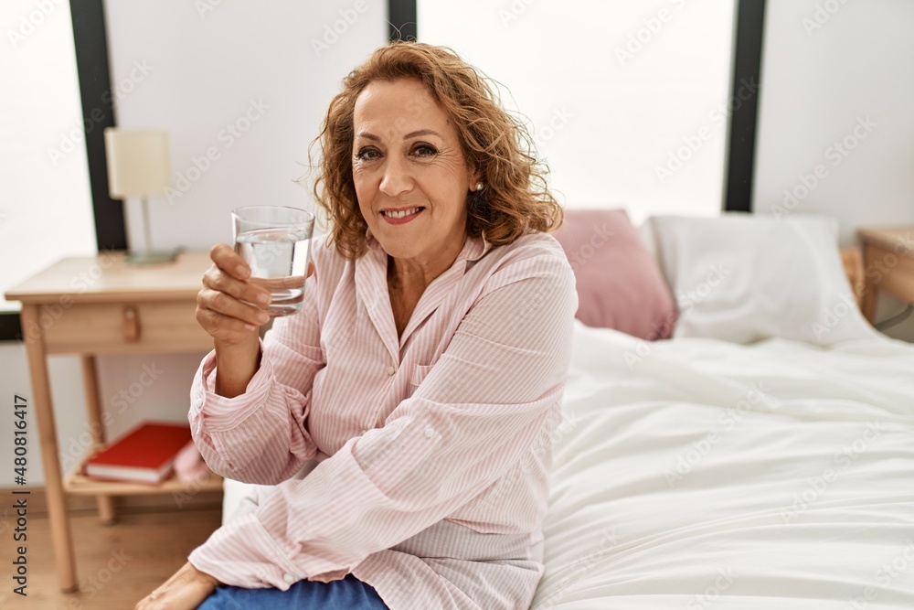 Middle age caucasian woman drinking water sitting on the bed at bedroom. Middle age caucasian woman drinking water sitting on the bed at bedroom.
