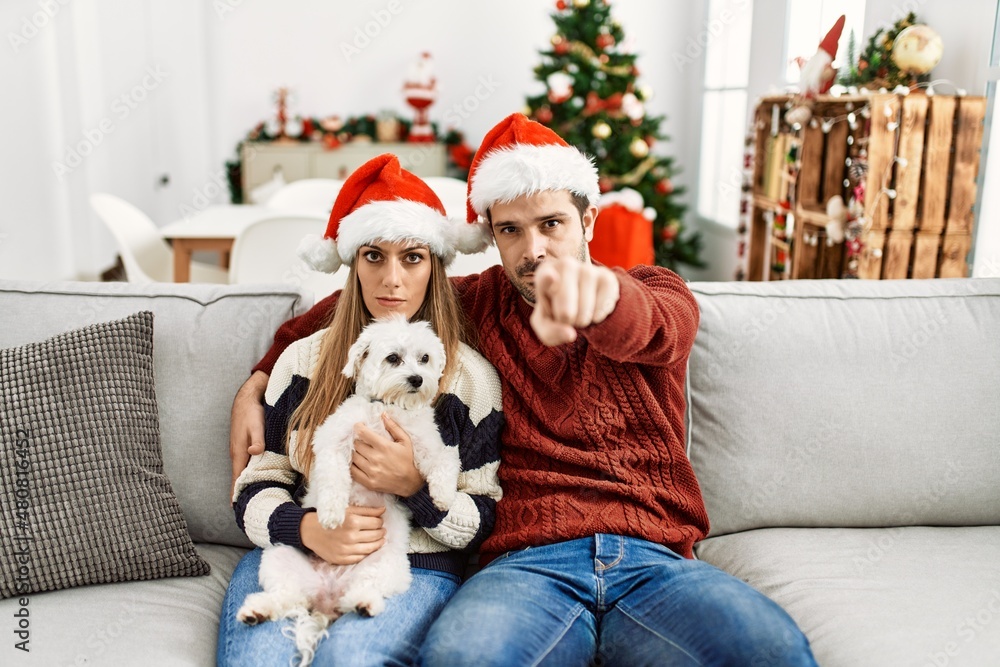 Young couple of wife and husband sitting on the sofa wearing christmas hat at home pointing with finger to the camera and to you, confident gesture looking serious Young couple of wife and husband sitting on the sofa wearing christmas hat at home pointing with finger to the camera and to you, confident gesture looking serious