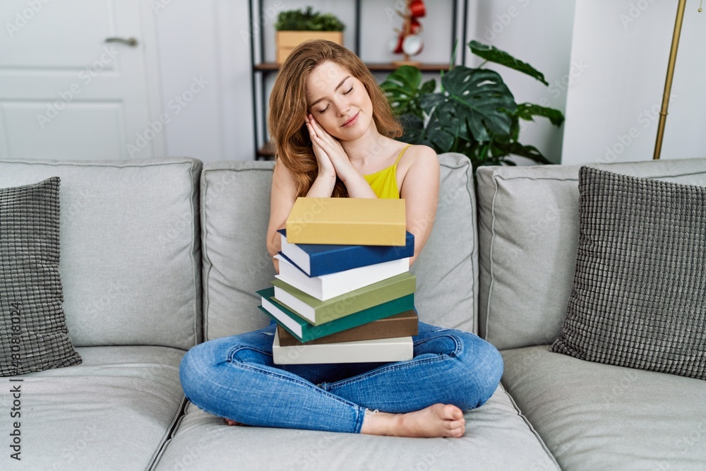 Young caucasian woman sitting on the sofa with books at home sleeping tired dreaming and posing with hands together while smiling with closed eyes. Young caucasian woman sitting on the sofa with books at home sleeping tired dreaming and posing with hands together while smiling with closed eyes.