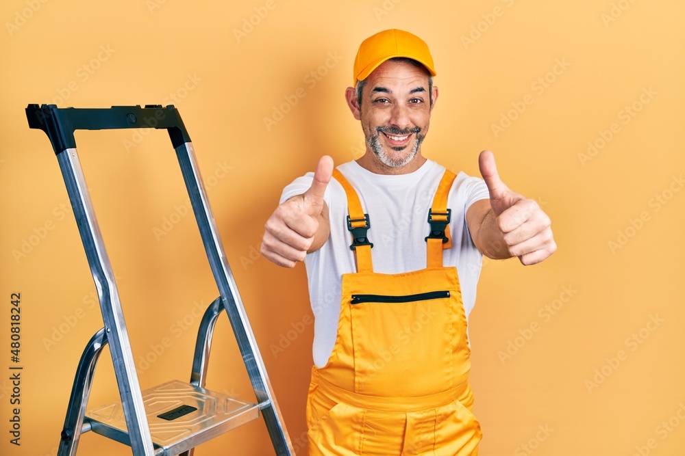 Handsome middle age man with grey hair holding ladder approving doing positive gesture with hand, thumbs up smiling and happy for success. winner gesture. Handsome middle age man with grey hair holding ladder approving doing positive gesture with hand, thumbs up smiling and happy for success. winner gesture.