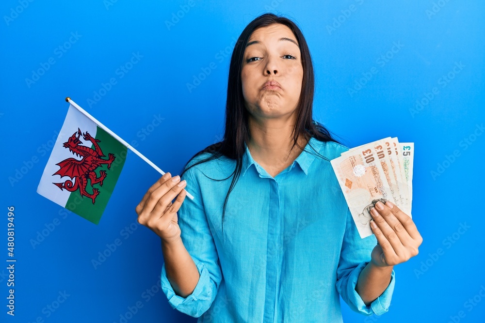 Young latin woman holding wales flag and pounds banknotes puffing cheeks with funny face. mouth inflated with air, catching air. Young latin woman holding wales flag and pounds banknotes puffing cheeks with funny face. mouth inflated with air, catching air.