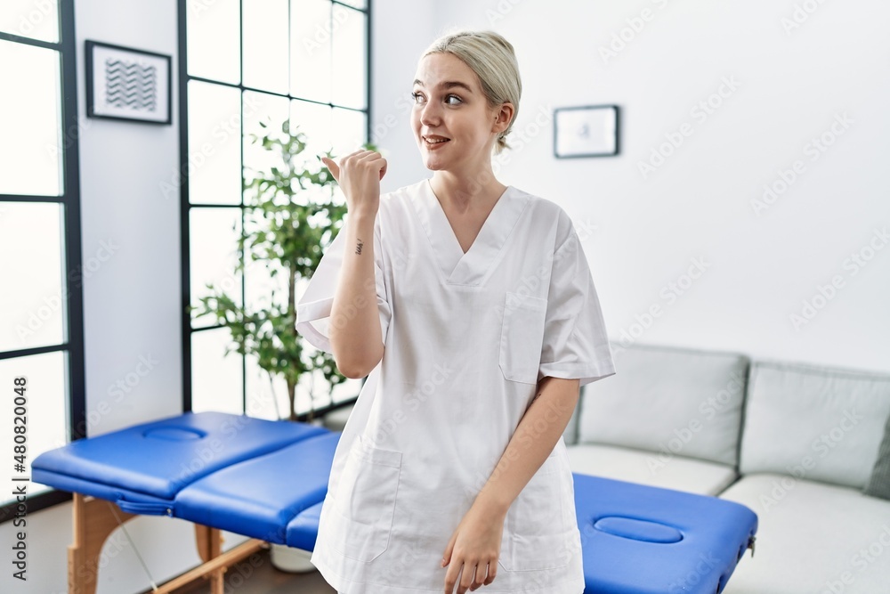 Young caucasian physiotherapy woman working at home smiling with happy face looking and pointing to the side with thumb up. Young caucasian physiotherapy woman working at home smiling with happy face looking and pointing to the side with thumb up.