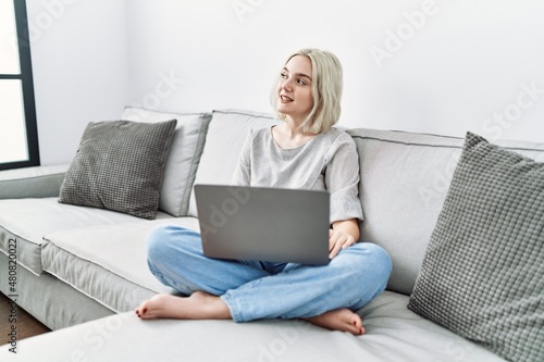 Young caucasian woman using laptop at home sitting on the sofa looking away to side with smile on face, natural expression. laughing confident.