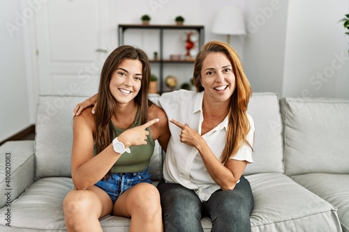 Mother and daughter together sitting on the sofa at home cheerful with a smile on face pointing with hand and finger up to the side with happy and natural expression