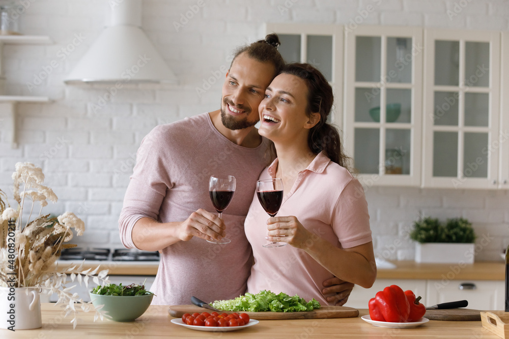 Happy bonding young man woman family couple with wineglasses in hands looking in distance, visualizing common future, imagining life together, dreaming enjoying romantic home date in modern kitchen.