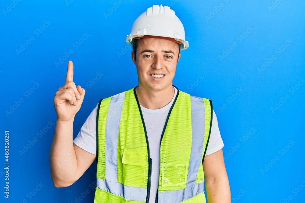Handsome young man wearing safety helmet and reflective jacket pointing finger up with successful idea. exited and happy. number one. Handsome young man wearing safety helmet and reflective jacket pointing finger up with successful idea. exited and happy. number one.