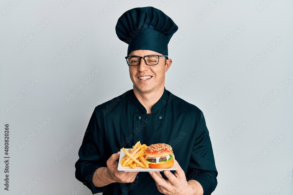 Handsome young man chef holding burger with fries winking looking at the camera with sexy expression, cheerful and happy face. Handsome young man chef holding burger with fries winking looking at the camera with sexy expression, cheerful and happy face.
