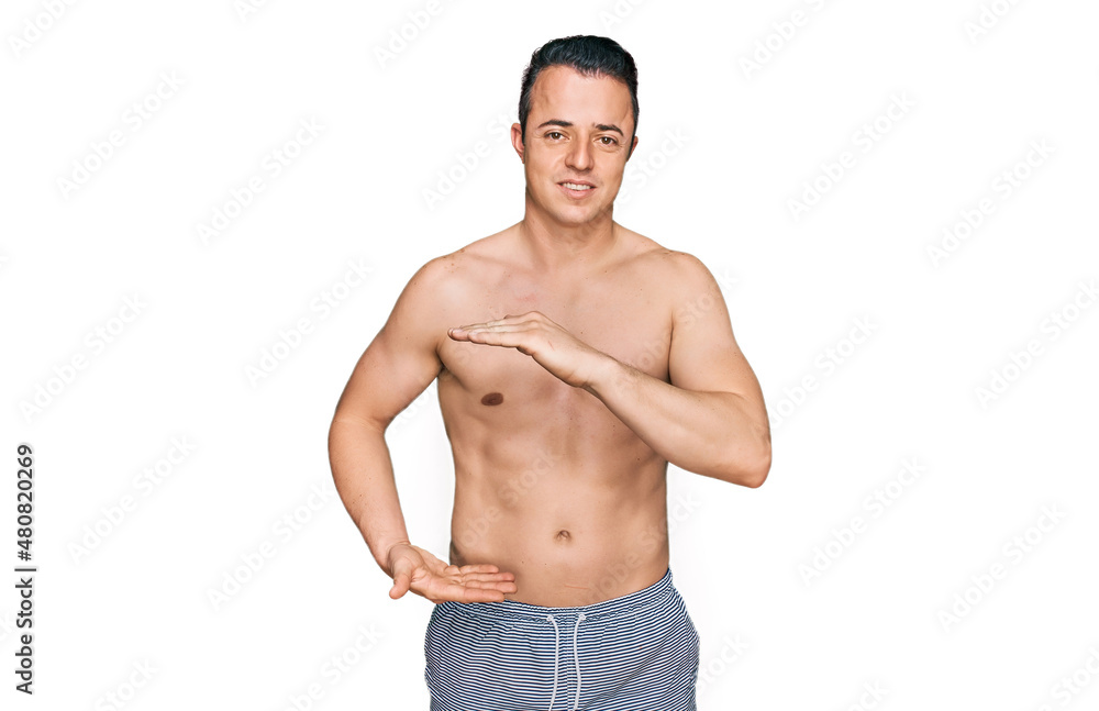 Fototapeta premium Handsome young man wearing swimwear shirtless gesturing with hands showing big and large size sign, measure symbol. smiling looking at the camera. measuring concept.
