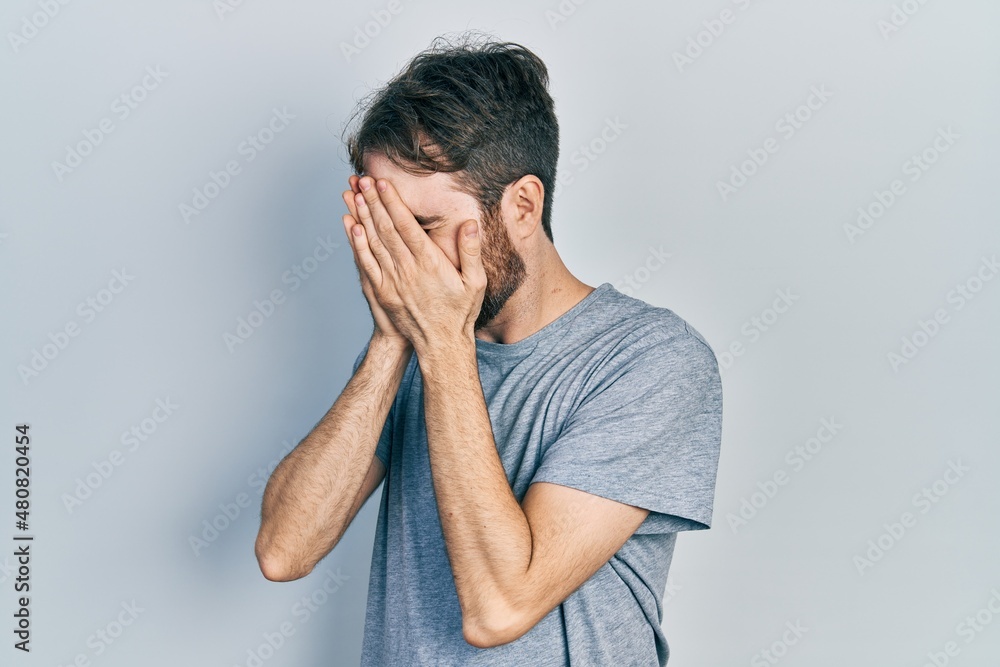 Caucasian man with beard wearing casual grey t shirt with sad expression covering face with hands while crying. depression concept. Caucasian man with beard wearing casual grey t shirt with sad expression covering face with hands while crying. depression concept.
