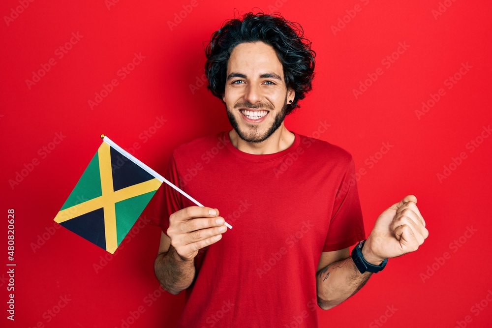 Handsome hispanic man holding jamaica flag screaming proud, celebrating victory and success very excited with raised arm Handsome hispanic man holding jamaica flag screaming proud, celebrating victory and success very excited with raised arm