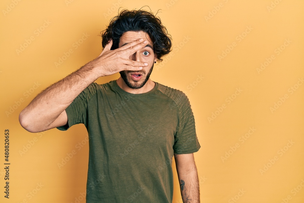 Handsome hispanic man wearing casual green t shirt peeking in shock covering face and eyes with hand, looking through fingers afraid Handsome hispanic man wearing casual green t shirt peeking in shock covering face and eyes with hand, looking through fingers afraid