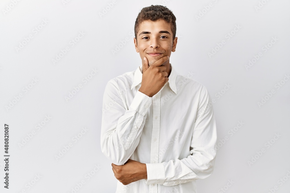 Young handsome hispanic man standing over isolated background looking confident at the camera smiling with crossed arms and hand raised on chin. thinking positive. Young handsome hispanic man standing over isolated background looking confident at the camera smiling with crossed arms and hand raised on chin. thinking positive.