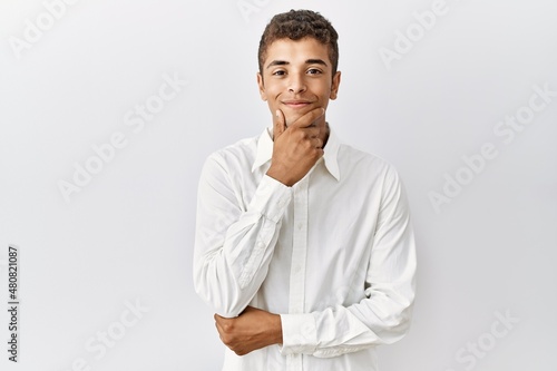 Young handsome hispanic man standing over isolated background looking confident at the camera smiling with crossed arms and hand raised on chin. thinking positive.