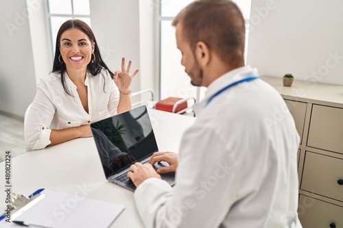 Young hispanic woman at the doctor doing ok sign with fingers  smiling friendly gesturing excellent symbol