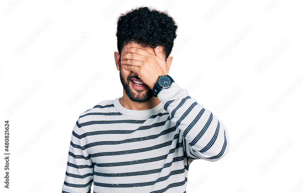 Young arab man with beard wearing casual striped sweater smiling and laughing with hand on face covering eyes for surprise. blind concept. Young arab man with beard wearing casual striped sweater smiling and laughing with hand on face covering eyes for surprise. blind concept.