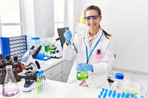 Young hispanic woman wearing scientist uniform using pipette and test tube working at laboratory