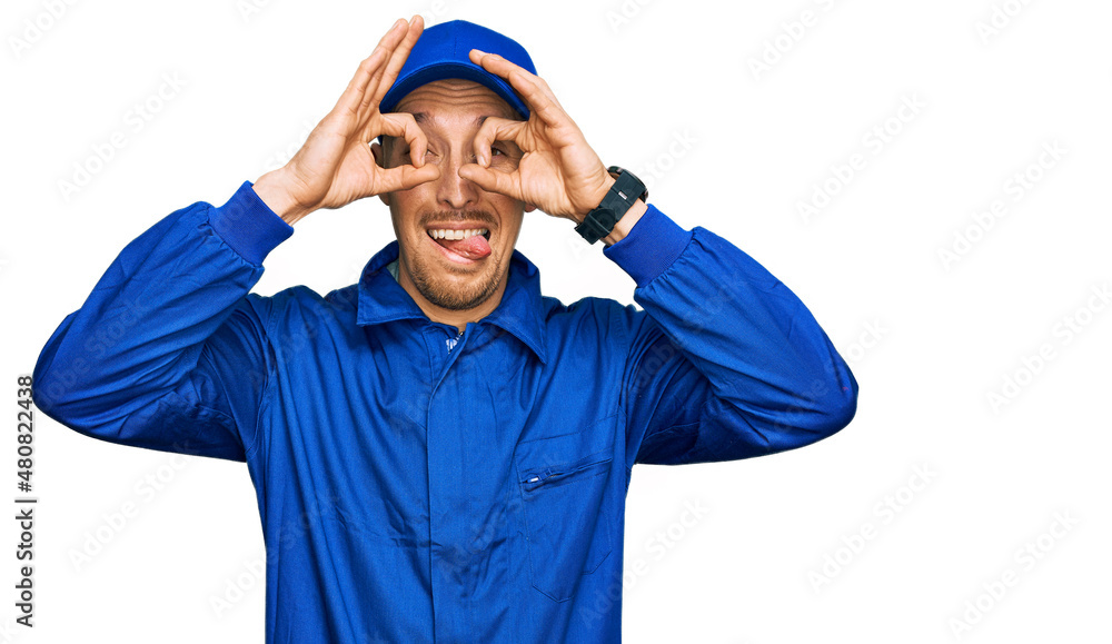 Naklejka premium Bald man with beard wearing builder jumpsuit uniform doing ok gesture like binoculars sticking tongue out, eyes looking through fingers. crazy expression.
