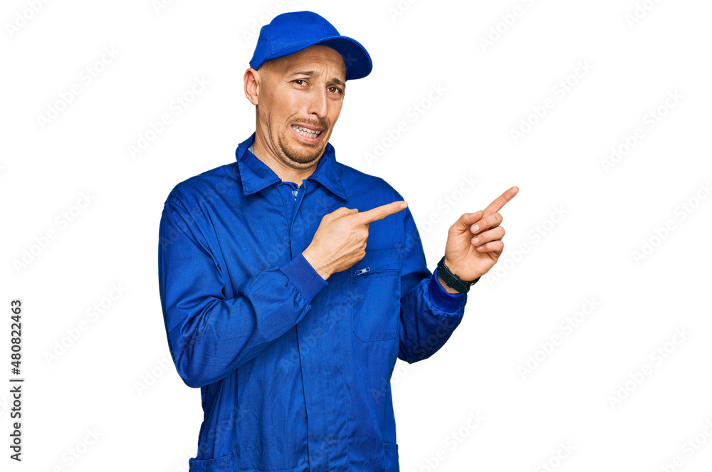 Bald man with beard wearing builder jumpsuit uniform pointing aside worried and nervous with both hands, concerned and surprised expression Bald man with beard wearing builder jumpsuit uniform pointing aside worried and nervous with both hands, concerned and surprised expression