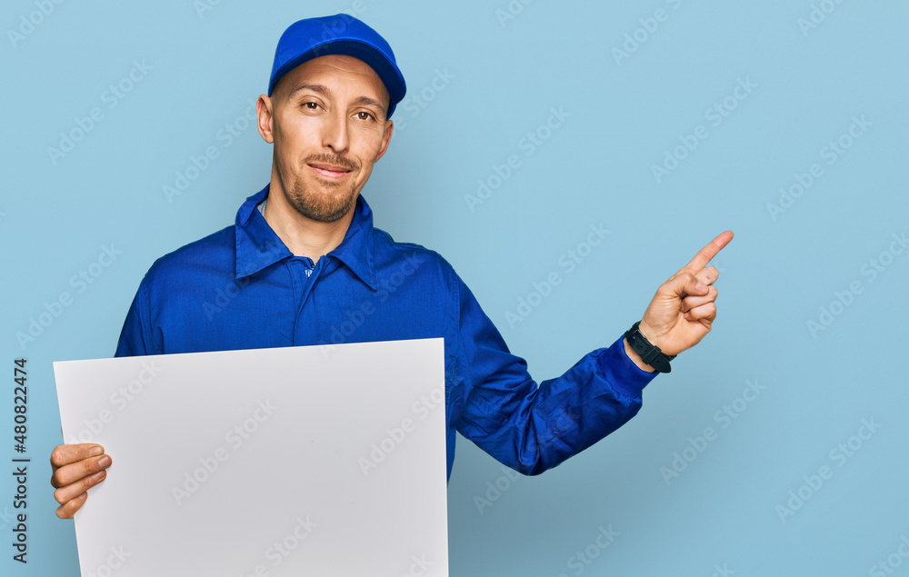 Bald man with beard wearing builder jumpsuit uniform holding empty banner smiling happy pointing with hand and finger to the side Bald man with beard wearing builder jumpsuit uniform holding empty banner smiling happy pointing with hand and finger to the side