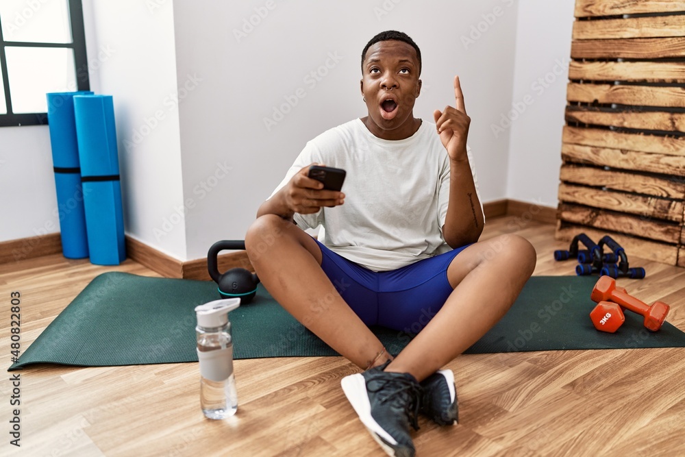 Young african man sitting on training mat at the gym using smartphone amazed and surprised looking up and pointing with fingers and raised arms. Young african man sitting on training mat at the gym using smartphone amazed and surprised looking up and pointing with fingers and raised arms.