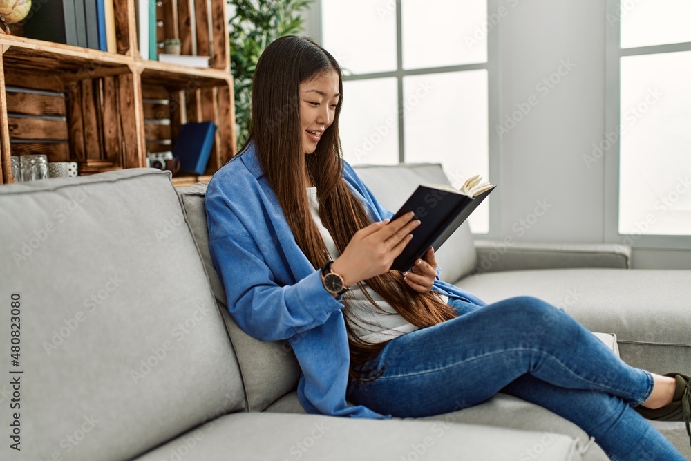 Young chinese girl reading book sitting on the sofa at home. Young chinese girl reading book sitting on the sofa at home.