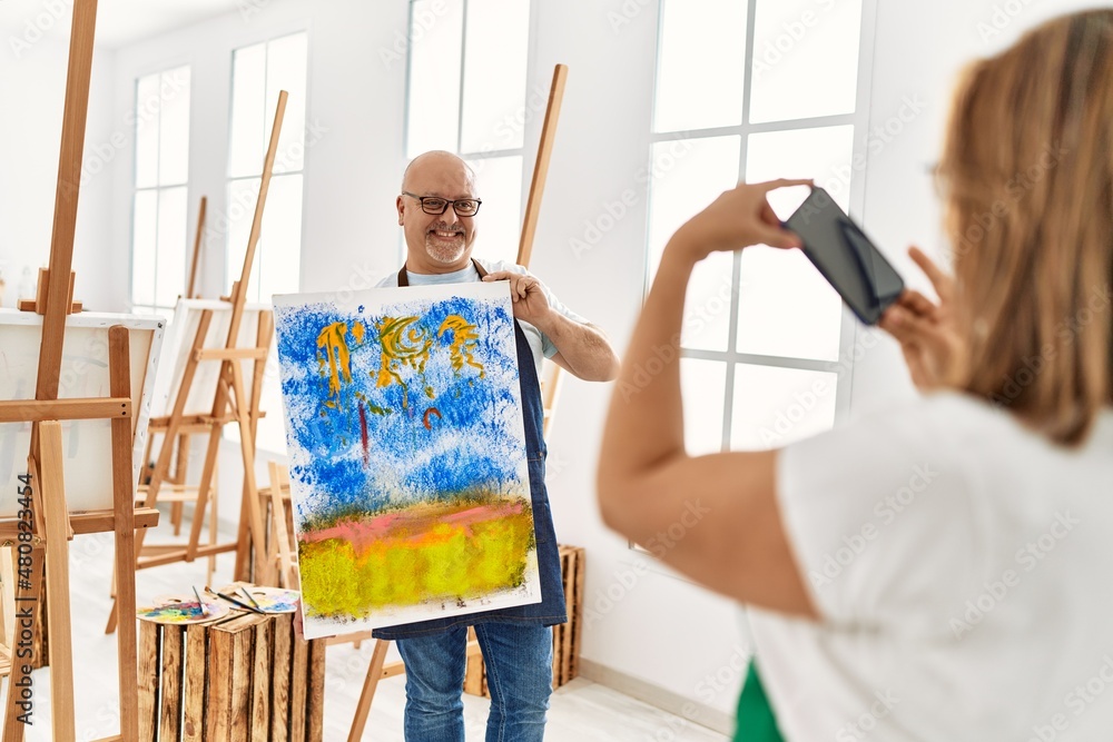 Middle age hispanic painter couple smiling happy making photo using smartphone at art studio.