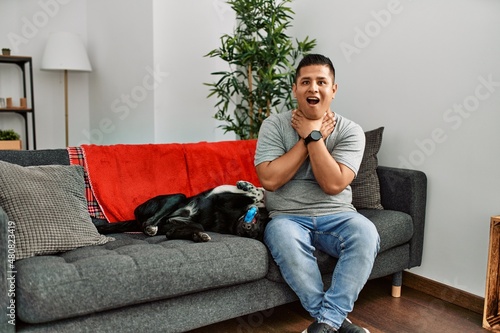 Young latin man and dog sitting on the sofa at home shouting suffocate because painful strangle. health problem. asphyxiate and suicide concept.