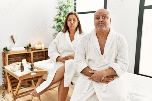 Middle age hispanic couple wearing bathrobe at wellness spa puffing cheeks with funny face. mouth inflated with air, crazy expression.