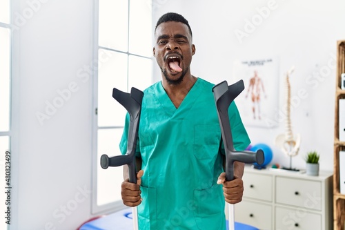 Young african physiotherapist man holding crutches sticking tongue out happy with funny expression Fotobehang