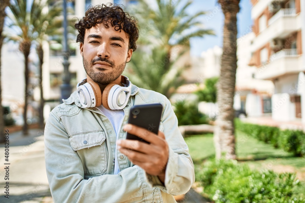 Fototapeta premium Handsome hispanic man with beard smiling happy outdoors on a sunny day wearing headphones listening to music with smartphone