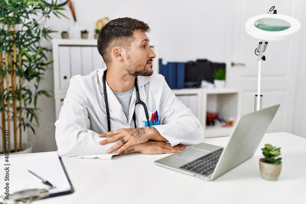 Young doctor working at the clinic using computer laptop looking to side, relax profile pose with natural face with confident smile.