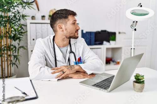 Young doctor working at the clinic using computer laptop looking to side, relax profile pose with natural face with confident smile.