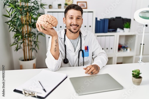 Young doctor holding brain at medical clinic looking positive and happy standing and smiling with a confident smile showing teeth