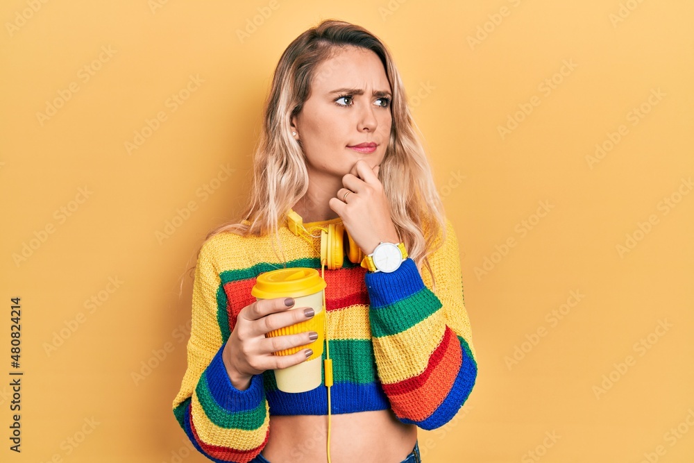 Beautiful young blonde woman drinking cup of coffee wearing headphones thinking worried about a question, concerned and nervous with hand on chin Beautiful young blonde woman drinking cup of coffee wearing headphones thinking worried about a question, concerned and nervous with hand on chin
