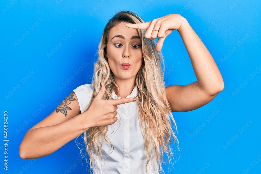 Beautiful young blonde woman doing picture frame gesture with hands making fish face with mouth and squinting eyes, crazy and comical.