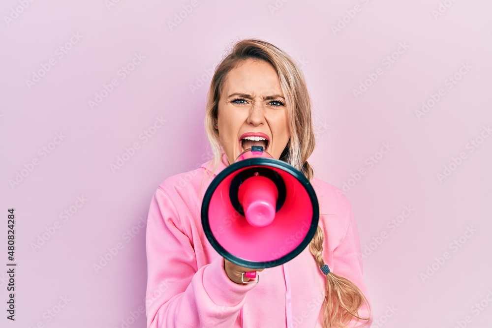 Fototapeta premium Young beautiful blonde caucasian woman shouthing and screaming through megaphone over isolated pink background