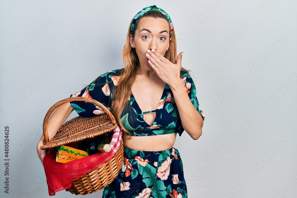Young caucasian woman holding picnic wicker basket covering mouth with hand, shocked and afraid for mistake. surprised expression