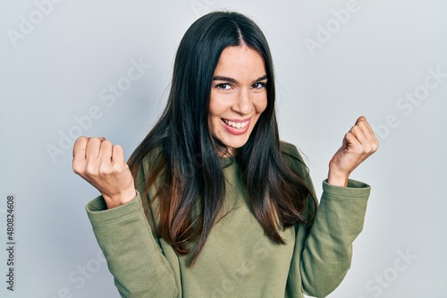 Young brunette woman wearing casual green sweater screaming proud, celebrating victory and success very excited with raised arms