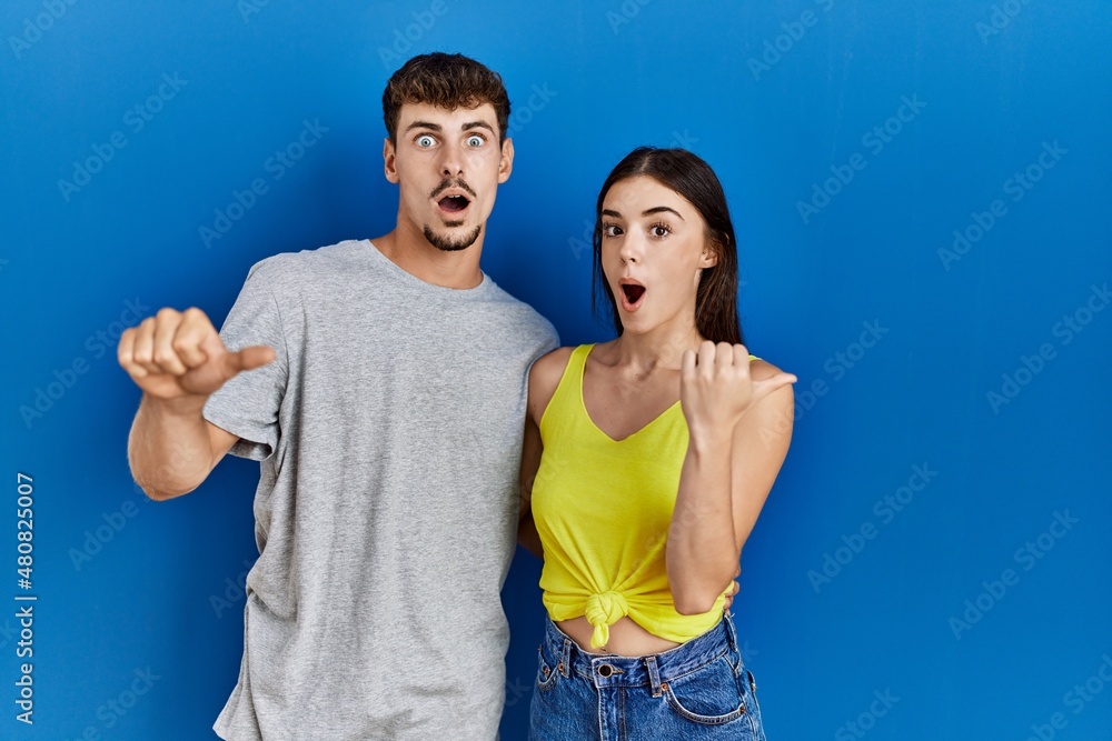 Young hispanic couple standing together over blue background surprised pointing with hand finger to the side, open mouth amazed expression. Young hispanic couple standing together over blue background surprised pointing with hand finger to the side, open mouth amazed expression.