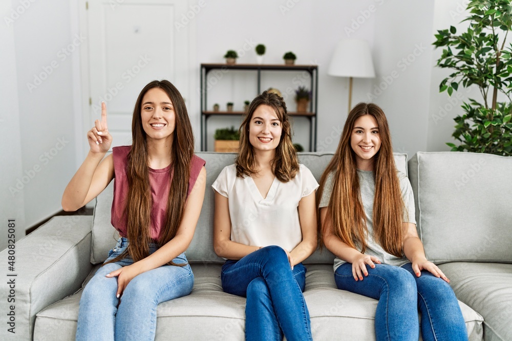 Group of three hispanic girls sitting on the sofa at home showing and pointing up with finger number one while smiling confident and happy. Group of three hispanic girls sitting on the sofa at home showing and pointing up with finger number one while smiling confident and happy.