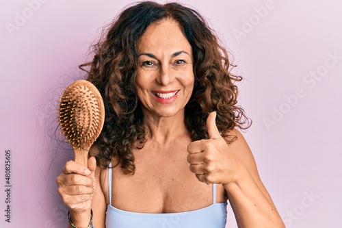Middle age hispanic woman using comb smiling happy and positive, thumb up doing excellent and approval sign
