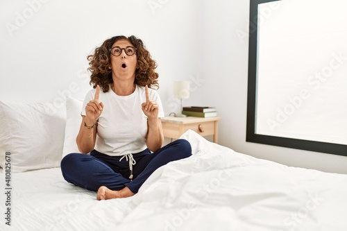 Wallpaper Mural Middle age hispanic woman sitting on the bed at home amazed and surprised looking up and pointing with fingers and raised arms. Torontodigital.ca