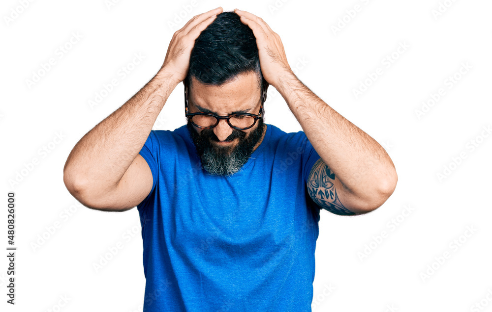 Hispanic man with beard wearing casual t shirt and glasses suffering from headache desperate and stressed because pain and migraine. hands on head. Hispanic man with beard wearing casual t shirt and glasses suffering from headache desperate and stressed because pain and migraine. hands on head.