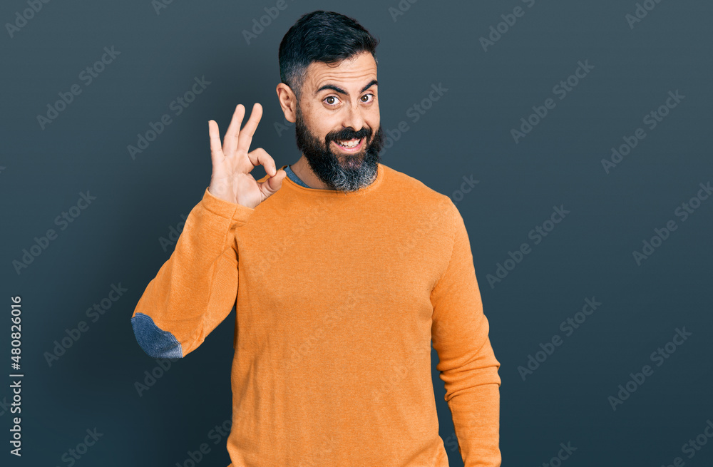 Hispanic man with beard wearing casual winter sweater smiling positive doing ok sign with hand and fingers. successful expression. Hispanic man with beard wearing casual winter sweater smiling positive doing ok sign with hand and fingers. successful expression.
