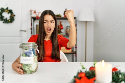 Young brunette woman on christmas holding charity jar with money at home annoyed and frustrated shouting with anger  yelling crazy with anger and hand raised