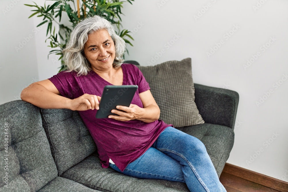 Obraz premium Middle age grey-haired woman using touchpad sitting on the sofa at home.