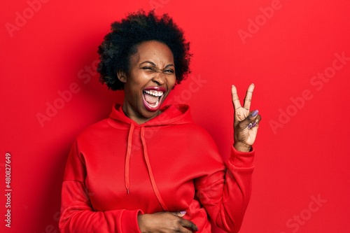 Young african american woman wearing casual sweatshirt smiling with happy face winking at the camera doing victory sign. number two.