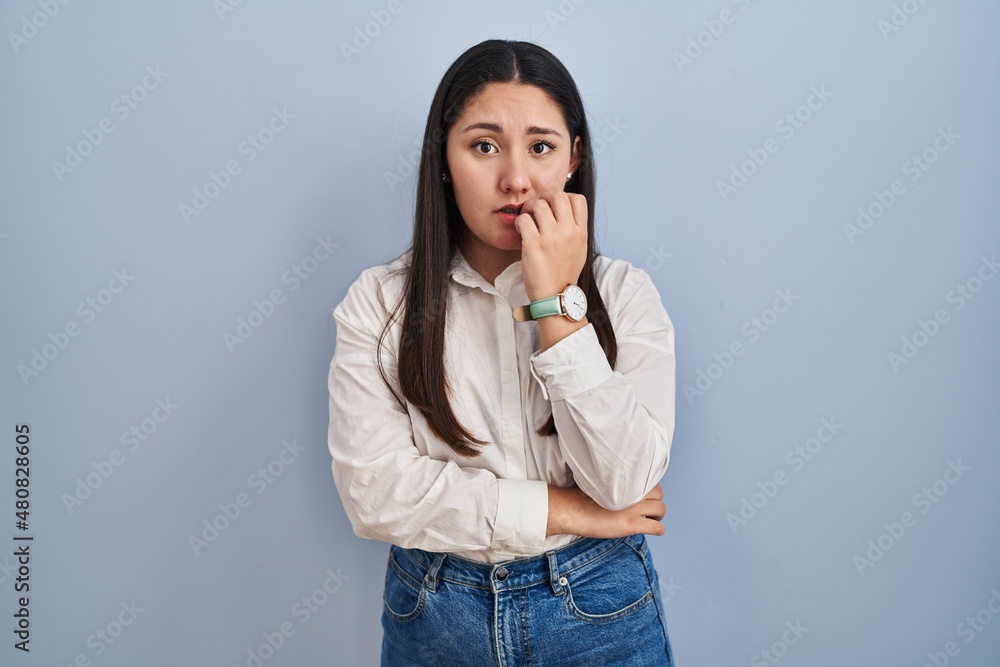 Young latin woman standing over blue background looking stressed and nervous with hands on mouth biting nails. anxiety problem. Young latin woman standing over blue background looking stressed and nervous with hands on mouth biting nails. anxiety problem.